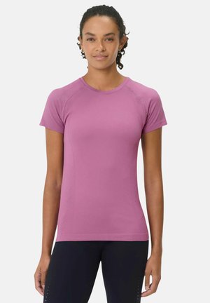 Vrouw met krullend haar draagt een nauwsluitend paars sportshirt met korte mouwen en zwarte leggings, staand tegen een eenvoudige lichte achtergrond.