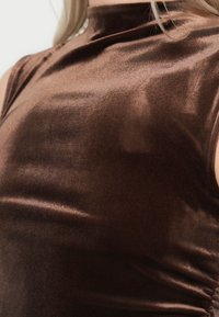 Καφέ τοπ με ψηλό λαιμό, κατασκευασμένο από γυαλιστερό, μεταξένιο ύφασμα με εφαρμοστό σχέδιο και διακριτική λάμψη. Οι σούρες στο πλάι προσθέτουν υφή και οπτικό ενδιαφέρον.