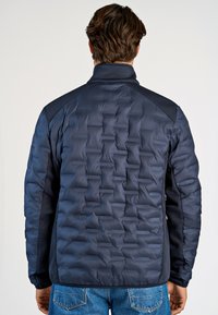 Marinblå puffjacka med quiltat mönster och meshdetaljer på axlarna. Texturerad tyg, hög krage och elastiska ärmslut.