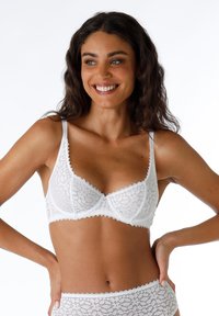 Bra bianco in pizzo con un delicato motivo floreale, bordi scallop, spalline regolabili e un trim decorativo.