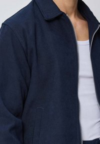 Veste zippée bleu marine avec un col pointu, arborant une texture lisse et des poches avant. Débardeur gris clair visible en dessous.