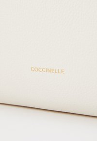 Bijela kožna površina s teksturiranim finišom koja sadrži zlatni urezani logo "Coccinelle".