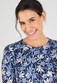 Marineblauwe bloemenblouse met een ronde hals; voorzien van grote lichtpaarse en roze bloemen met groene bladeren; zachte stof, subtiele glans en plooien op de schouders.