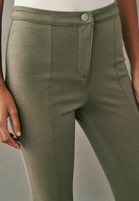 Pantalones elásticos de color verde oliva con una textura suave, que presentan un cierre con botón en la parte delantera y bolsillos, destacados por un sutil diseño de costuras.