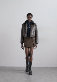 Giubbotto corto in pelle marrone con colletto di pelliccia, abbinato a un polo blu scuro e una mini gonna marrone. Stivali neri fino al ginocchio completano l'outfit.