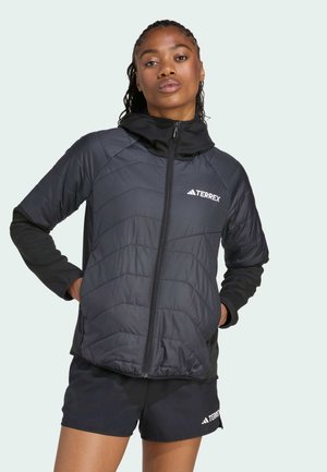 A young person poses confidently in a black TERREX jacket and matching shorts, showcasing a sporty look against a light background.