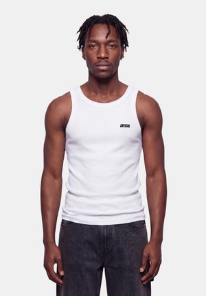 Mężczyzna z warkoczami, ubrany w biały tank top z napisem „CRYSTAL” i czarne jeansy, stoi na tle jednolitego jasnego tła.