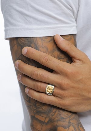 Hand mit einem silbernen Ring mit einem gehämmerten goldenen Quadrat ruht auf einem tätowierten Unterarm, teilweise bedeckt von einem weißen Ärmel.