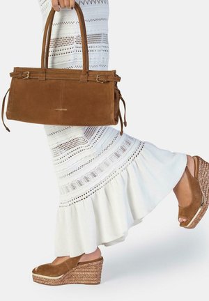 Donna in vestito bianco all'uncinetto tiene una borsa in camoscio marrone e indossa sandali con zeppa marroni, un piede sollevato dietro di lei.