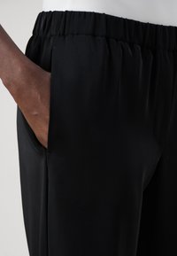 Svarta byxor med elastiskt midjeband och en slät yta, med sidfickor och en avslappnad passform. Rena linjer och minimala design detaljer.