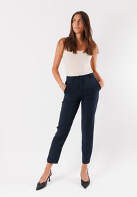 Pantaloni navali sartoriali con vita alta, tasche laterali, pieghe e una finitura in tessuto liscia, abbinati a una canotta aderente di colore crema e tacchi neri.