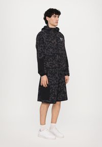 Black lightweight jacket and matching shorts in a camouflage pattern, featuring an adjustable hood and multiple pockets. White sneakers.