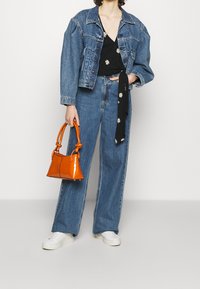 Giacca di jeans con maniche a sbuffo, top nero con decorazioni a margherita, jeans a gamba larga, borsa lucida arancione e sneakers bianche.