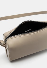 Sac à main en cuir beige avec une finition lisse, fermeture éclair et étiquette intérieure. Il dispose d'une bandoulière unique et d'une forme structurée.