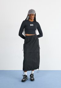 Fekete, hosszú ujjú cropped top fehér logóval, fekete midi szoknyával párosítva, amelyen cargo zsebek és állítható húzózsinórok találhatók. Sötét sportcipő.