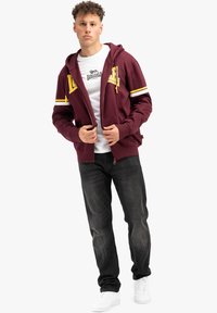 Burgundská mikina so zipsom a žltými akcentami, dve predné vrecká a kapucňa na šnúrku, nosená cez bielu tričko a čierne džínsy.