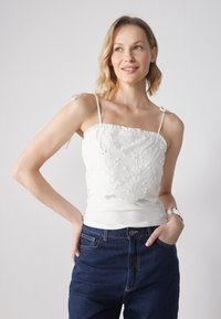 Hvit cami med blomster broderi, volang kant på toppen og justerbare stropper, sammen med mørkeblå høy midje jeans. Klassisk design.
