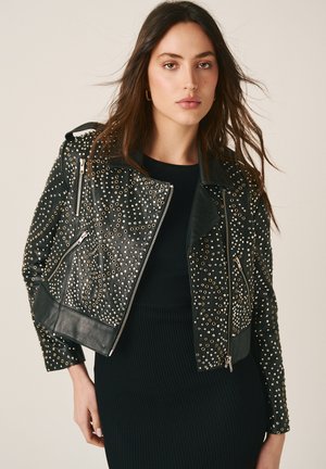 Jonge vrouw die een zwarte leren jas met studs draagt over een zwarte geribbelde jurk, tegen een effen beige achtergrond.