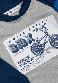 Pelēks un zils treniņtērps ar BMX velosipēda grafiku. Monstrē liela balta plāksteru ar zilu tekstu un ilustrācijām, kokvilnas audumu un sportisku dizainu.