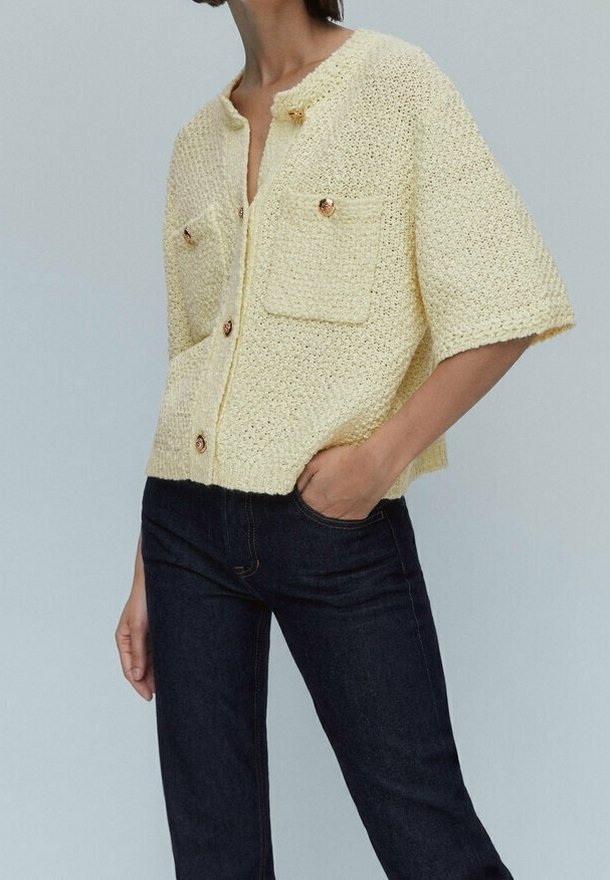 Krótki rękaw, zapinany kardigan w miękkim żółtym dzianinie, z dwiema przednimi kieszeniami i ozdobnymi guzikami, w połączeniu z ciemnymi dżinsami.