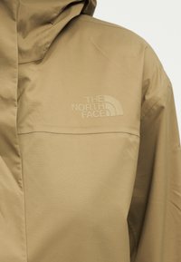 Rudos spalvos nepralaidus vandeniui striukė su kapišonu, ant krūtinės iškilusiu The North Face logotipu.