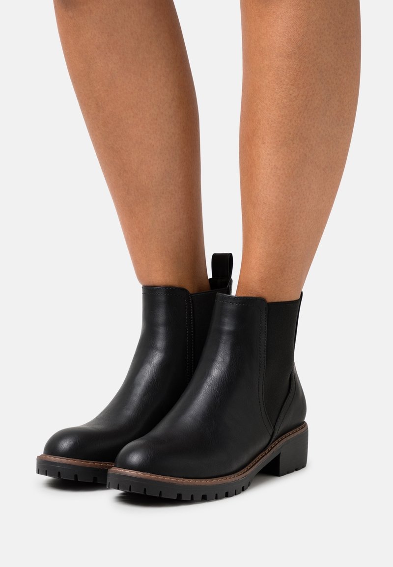 Bottines en cuir noir avec languettes, talons épais et semelles crantées portées sur des jambes nues sur un fond blanc uni.