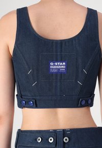 Denimski kratki top v temno modri barvi z квадратным kepkom, ki vsebuje napis "G-STAR ORIGINALS RAW DENIM." Vključuje gumbne detajle in bele šive.