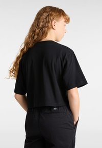 Černé zkrácené tričko s krátkým rukávem, vyrobené z bavlny, s minimalistickým designem a štítkem na zadní straně. Nošeno s černými kalhotami.