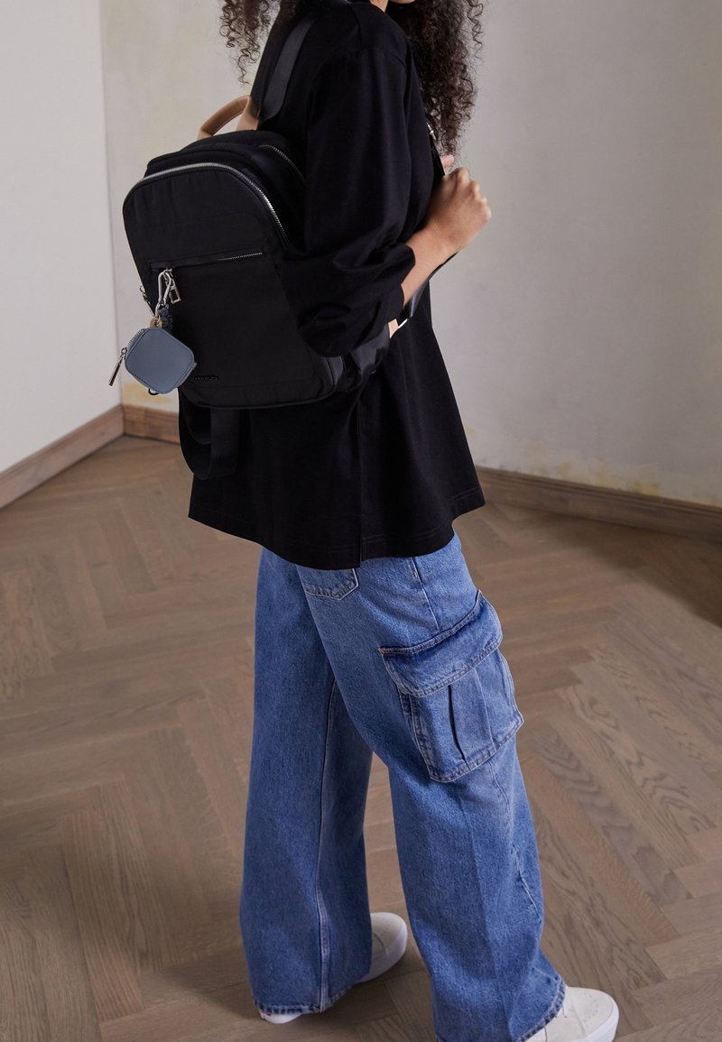 Personne portant un haut noir et un jean cargo bleu, portant un sac à dos noir avec une petite pochette grise attachée.
