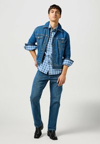 Giacca di jeans con chiusura a bottoni, tasche sul petto e maniche arrotolate; sovrapposta a una camicia a quadri blu. Abbinata a jeans blu a gamba dritta.
