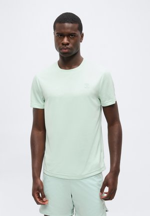 Uomo in piedi di fronte, indossa una camicia verde chiaro a maniche corte e pantaloncini abbinati su uno sfondo bianco semplice.