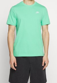 Muž v mátově zeleném tričku Nike s malým bílým logem na hrudi a černých šortkách, stojící na jednolitém pozadí.
