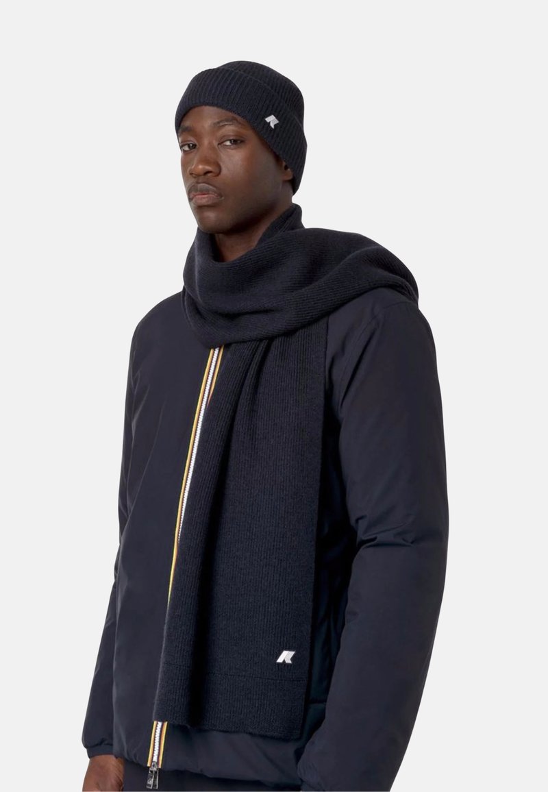 Bonnet en tricot bleu marine et écharpe assortie, présentant une texture côtelée, drapée avec élégance. L'écharpe comporte un détail de logo subtil et des lignes épurées.