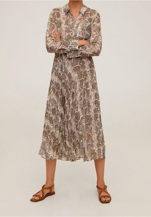 Woman wearing long-sleeve, buttoned, snake print midi dress and brown open-toe flat sandals, standing with arms crossed against plain background.