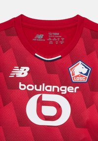 Sarkana bērnu futbolista krekls ar apaļu kaklu, struktūru rakstu, New Balance logo un kluba emblēmu. Izmēra birka: 12/18M.