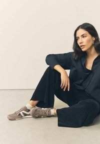 Graue und beige Sneaker mit einem strukturierten Muster, kombiniert mit schwarzen weit geschnittenen Hosen und einem dunklen Hemd mit Knöpfen, auf einer neutralen Oberfläche sitzend.