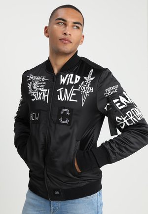 Giacca bomber nera con stampe grafiche bianche che includono testo e simboli. Caratterizzata da chiusura con zip, polsini a costine e tasche. Texture satinata liscia.