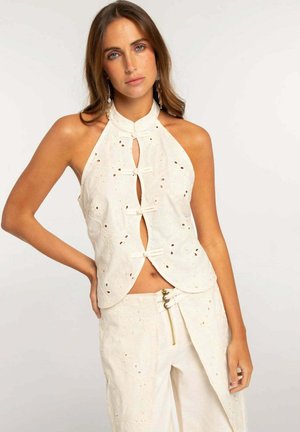 Mujer con cabello castaño claro lleva un top halter blanco sin mangas bordado y pantalones a juego de talle alto con cremallera y detalle de hebilla.