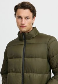 Grønn pufferjakke med horisontal quiltedesign, høy krage og frontglidelås. Glatt, lett materiale med matt overflate.