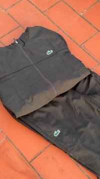 Conjunto de treino preto feito de material leve, composto por uma jaqueta com fecho éclair e calças combinando. Inclui pequenos acentos de logotipo verde.