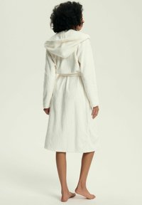 Szlafrok z kapturem w jasno-kremowym kolorze, wykonany z miękkiego, prążkowanego materiału, z paskiem w talii i długimi rękawami. Długość do kolan, o luźnym kroju.