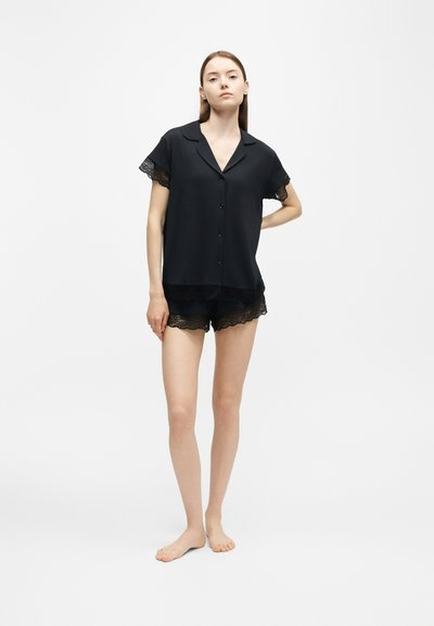 Jeune femme debout pieds nus portant un haut noir à manches courtes boutonné et un short assorti bordé de dentelle sur un fond blanc uni.