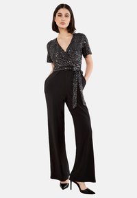 Vrouw in een zwart glinsterend wikkeltop met korte mouwen, gecombineerd met hooggeplaatste, wijdlopende zwarte broek en zwarte puntige hakken.