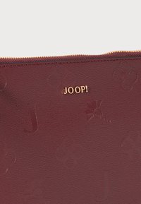 Bolsa de couro cor burgundy com padrão floral em relevo, logótipo "JOOP!" em tom dourado e fecho zip no topo.