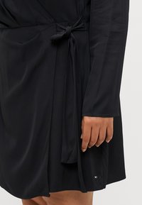 Vestito nero avvolgente con una cintura in vita, maniche lunghe, tessuto morbido e un sottile accento del logo vicino all'orlo. Texture liscia in tutto il vestito.