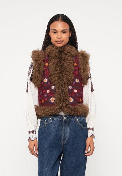 Nepvachtvest in bruin, met een bloempatronen bordeauxrode voorpaneel. Heeft een hoge kraag en een gestructureerde rand, gedragen over een witte blouse.
