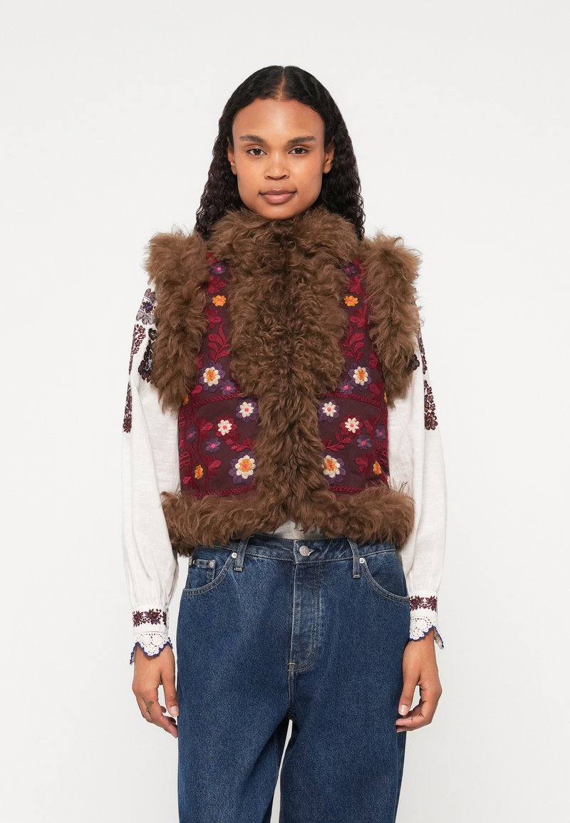 Hnedá umelá kožušina s kvetinovým vzorom na burgundskom prednom paneli. Má vysoký golier a textúrovaný lem, nosená cez bielu blúzku.