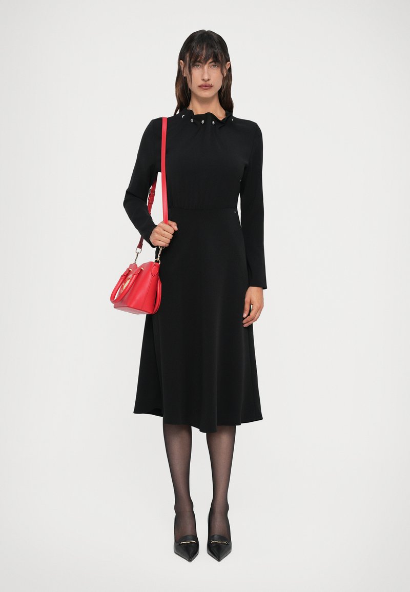 Abito lungo nero a maniche lunghe con busto aderente e gonna fluente. Presenta un colletto plissettato e una borsa rossa. Gambe adornate con calze trasparenti.