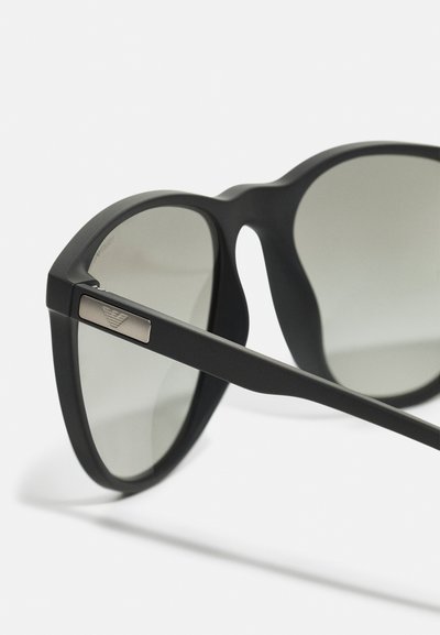 Occhiali da sole neri con lenti grigie sfumate, caratterizzati da una finitura opaca, una montatura ampia e un'applicazione metallica sulla stecca.