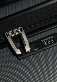 Superficie della valigia nera texturizzata con cerniera doppia argentata e lucchetto approvato TSA dotato di un foro per chiave circolare e un quadrante per combinazione a tre cifre.